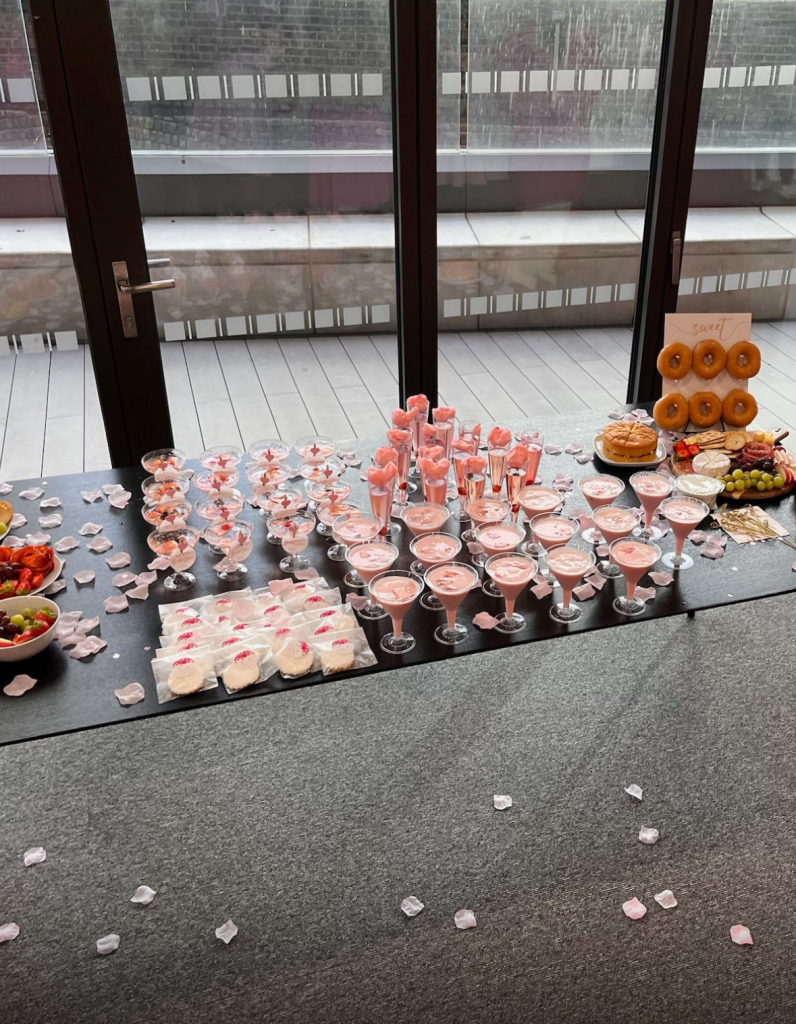 A beautifully arranged dessert table featuring pink cocktails, sweets, fruits, and donuts, adorned with rose petals.